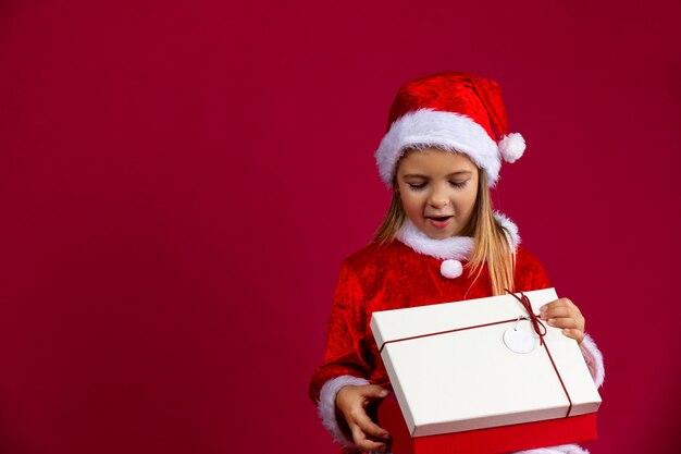 Bambina in un abito di Natale che indossa un cappello da Babbo Natale con sorpresa apre un regalo su un muro rosso con uno spazio vuoto laterale.