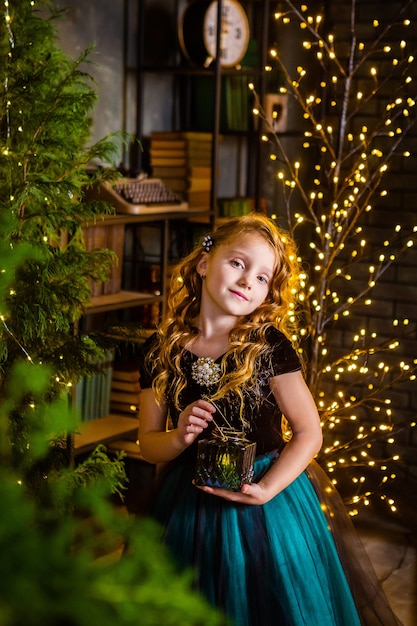 Bambina in un abito festivo ans lunghi capelli ricci con la candela in mano, albero di Natale sullo sfondo. Concetto di Natale e miracoli, decorazioni di Capodanno.