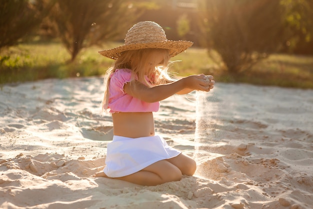 bambina in un cappello di paglia si siede su una spiaggia di sabbia in estate