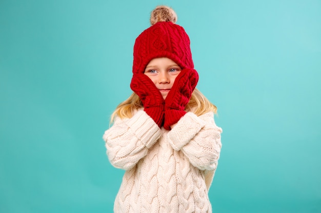 bambina in un cappello rosso lavorato a maglia, guanti e un maglione bianco sorride su una parete blu