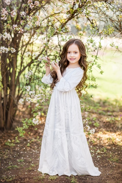 Bambina in un lungo abito bianco in un giardino primaverile. Un bambino vicino a un albero in fiore