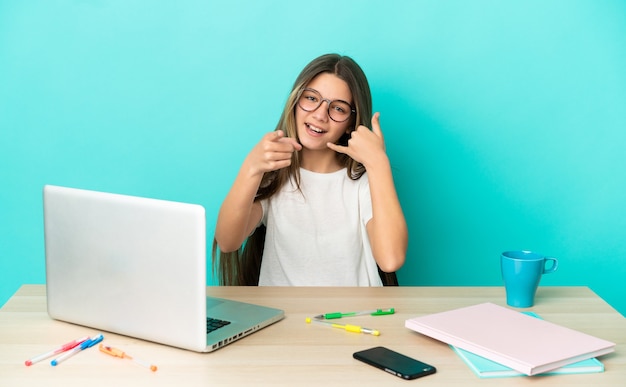 Bambina in un tavolo con un computer portatile su sfondo blu isolato che fa il gesto del telefono e indica la parte anteriore