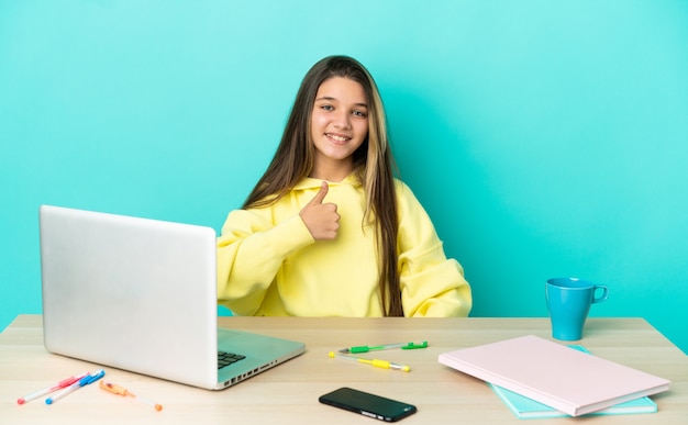 Bambina in un tavolo con un computer portatile su sfondo blu isolato che fa un gesto di pollice in alto