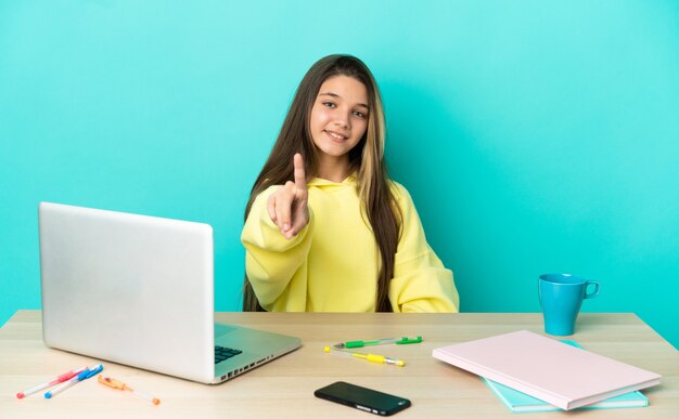 Bambina in un tavolo con un computer portatile su sfondo blu isolato che mostra e solleva un dito