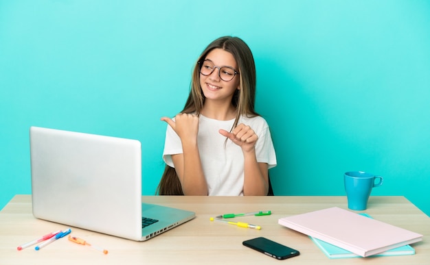 Bambina in un tavolo con un computer portatile su sfondo blu isolato che punta al lato per presentare un prodotto