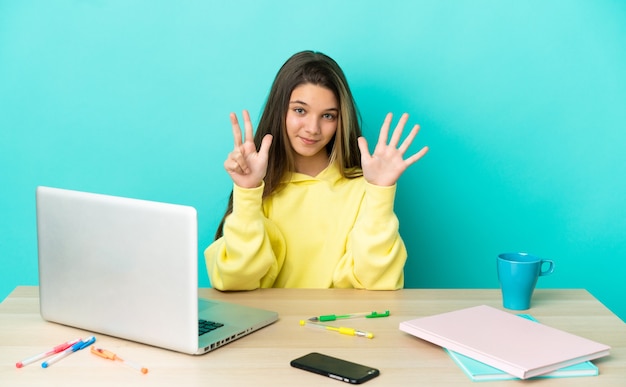 Bambina in un tavolo con un computer portatile su sfondo blu isolato contando otto con le dita