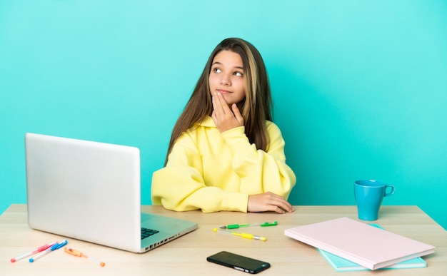 Bambina in un tavolo con un computer portatile su sfondo blu isolato guardando in alto mentre sorride