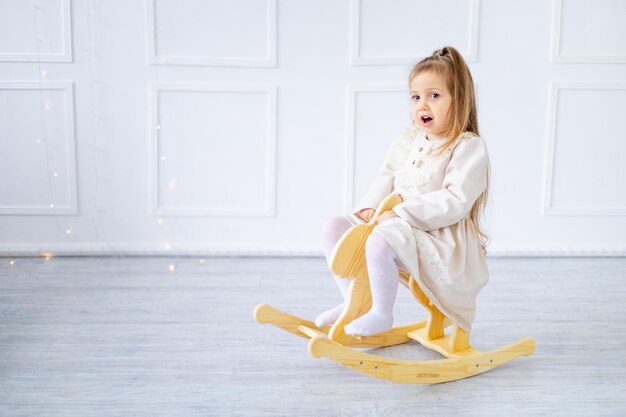 Bambina in un vestito di cotone che oscilla o gioca su un cavallo di legno in una stanza luminosa, spazio per il testo