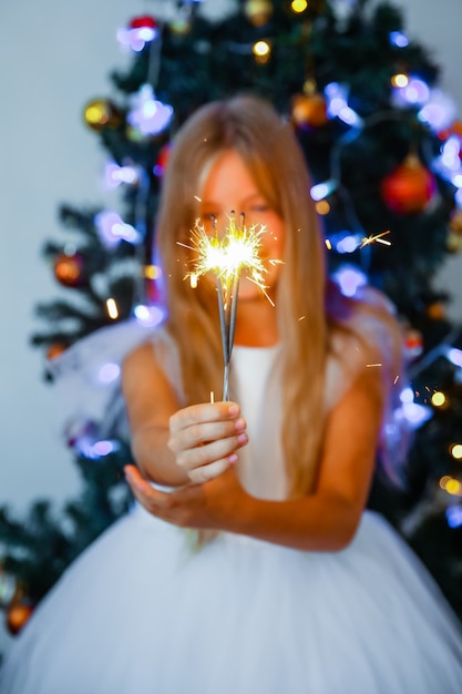 Bambina in un vestito festivo che tiene le stelle filanti sullo sfondo di un albero di Natale