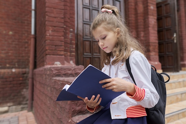 Bambina in uniforme con zaino seduto nel taccuino di lettura cortile della scuola, copia spazio. Ritorno a scuola, inizio delle lezioni, educazione, conoscenza, lezioni, concetto di bambini