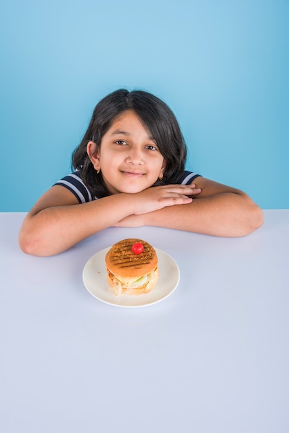 Bambina indiana o asiatica carina che mangia hamburger, sandwich o pizza gustosi in un piatto o in una scatola. In piedi isolato su sfondo blu o giallo.