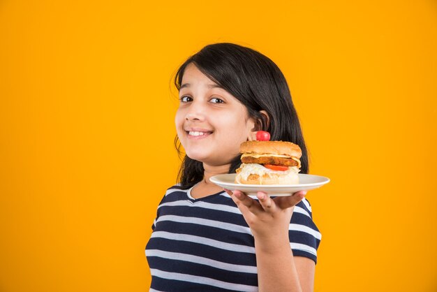 Bambina indiana o asiatica carina che mangia hamburger, sandwich o pizza gustosi in un piatto o in una scatola. In piedi isolato su sfondo blu o giallo.