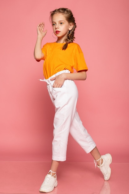 Bambina integrale sulla parete rosa dello studio. indossa una maglietta arancione e pantaloni bianchi e scarpe da ginnastica bianche.