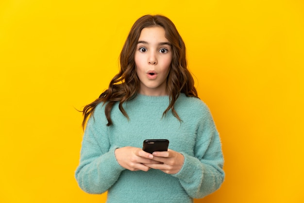 Bambina isolata sul muro giallo guardando la telecamera mentre si utilizza il cellulare con espressione sorpresa