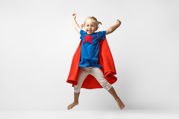 Bambina molto eccitata vestita da eroe che salta lungo il muro bianco.