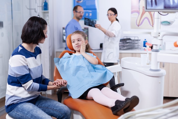 Bambina nell'ufficio del dentista che mostra al genitore dove il dente fa male toccando il viso con un'espressione dolorosa. Bambino con sua madre durante il controllo dei denti con stomatolog seduto su una sedia.