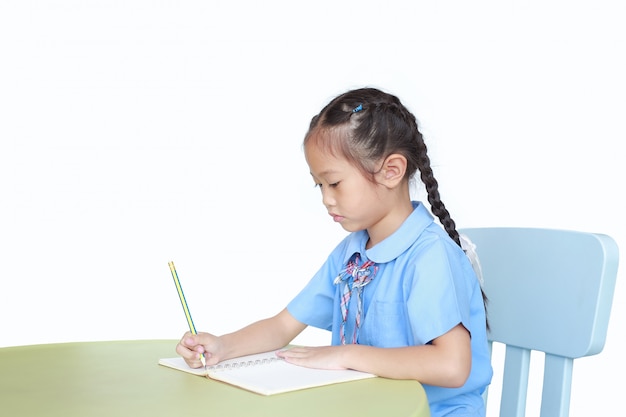 Bambina nella scrittura dell'uniforme scolastico sul libro allo scrittorio su fondo bianco