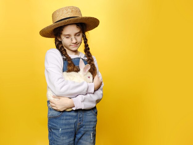 Bambina pensierosa in jeans e cappello di paglia e occhiali da sole rossi in possesso di un vero coniglio isolato sfondo giallo