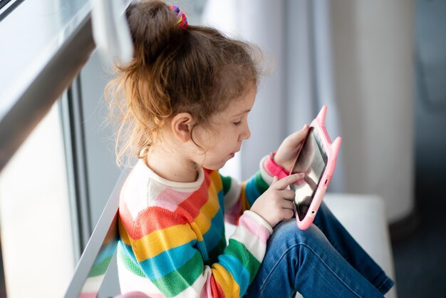 Bambina positiva, tenendo il computer tablet nelle sue mani. Pubblicità nuovo gadget, app educativa. La ragazza si siede vicino alla finestra. Indossa una maglietta colorata. Giochi. Formazione scolastica. I bambini usano tec