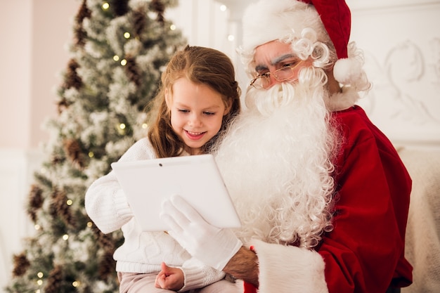Bambina seduta con Babbo Natale con tablet. Sulla poltrona di casa in soggiorno