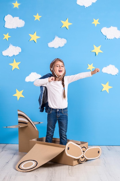 Bambina sognatrice che gioca con un aeroplano di cartone in studio con sfondo di cielo azzurro e nuvole bianche.