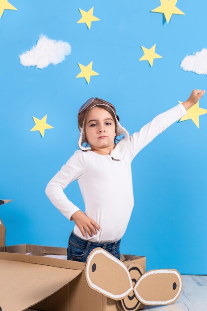 Bambina sognatrice che gioca con un aeroplano di cartone in studio con sfondo di cielo azzurro e nuvole bianche.