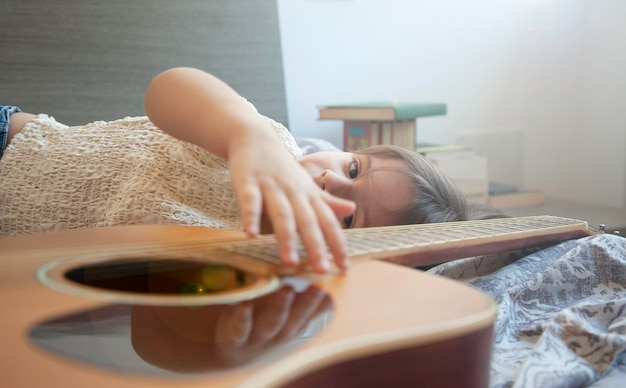 Bambina sognatrice con bellissimi occhi sdraiata sul letto con la sua chitarra acustica Sta toccando le corde della chitarra Il sogno è diventare un chitarrista in futuro Concetto di amante della musica Essere un sognatore