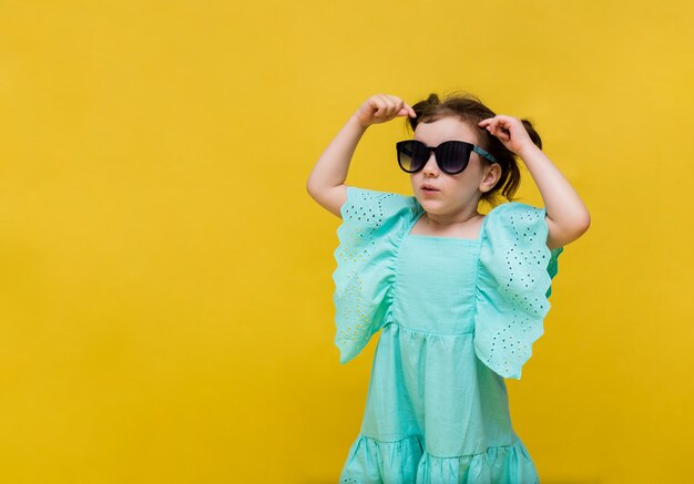 Bambina sorpresa in un vestito blu e occhiali da sole su uno sfondo giallo con spazio per il testo