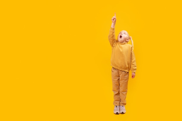 Bambina sorpresa punta il dito verso l'alto Ritratto a figura intera di una ragazza in età prescolare su sfondo giallo brillante Copia spazio spazio per il testo