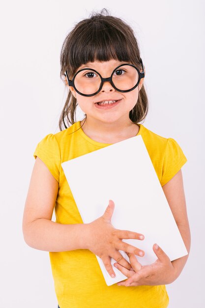 Bambina sorridente che indossa una maglietta gialla e occhiali neri rotondi che tiene un libro in mano su sfondo bianco