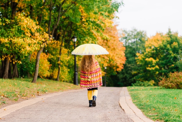 Bambina sorridente con l'ombrello in un impermeabile e stivali all'aperto