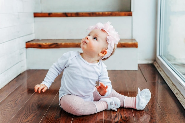 Bambina sorridente di un anno che indossa una corona di primavera situata sul pavimento in un soggiorno luminoso vicino alla finestra e gioca con i fiori di gerbera Bambino felice che gioca a casa Concetto di infanzia