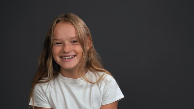 Bambina sorridente felice con capelli biondi in piedi guardando la parte anteriore che indossa la maglietta bianca isolata sul muro grigio scuro