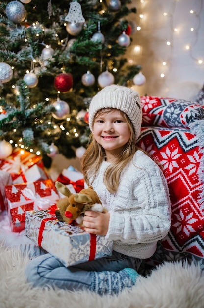 Bambina sorridente felice in cappello bianco che tiene scatole regalo di Natale mentre era seduto sul pavimento.