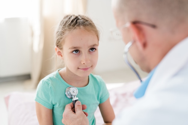 Bambina sorridente guardando il medico mentre lui esamina il suo petto con lo stetoscopio in ospedale