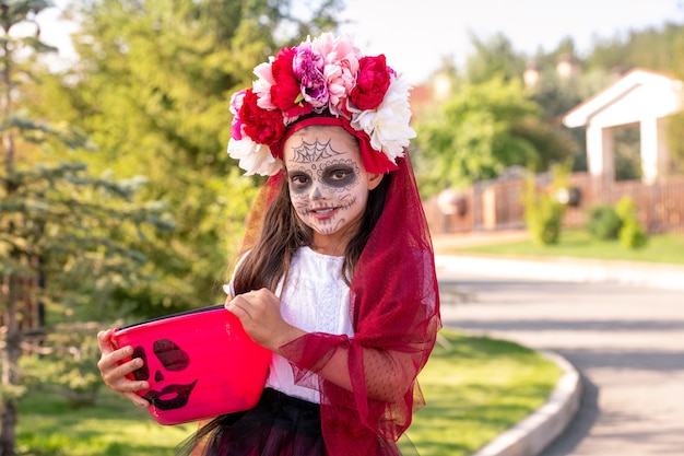 Bambina sorridente in costume da strega di Halloween che tiene in mano un cesto di plastica rosa o un secchio pieno di dolcetti mentre si trova davanti alla telecamera