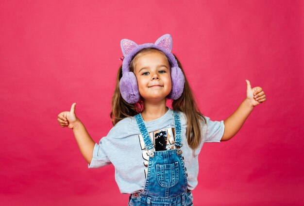 Bambina sorridente sveglia che si diverte con le cuffie di pelliccia su uno sfondo rosa.