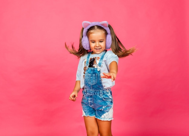 Bambina sorridente sveglia che si diverte con le cuffie di pelliccia su uno sfondo rosa.