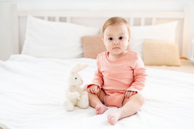 Bambina sul letto che gioca con un peluche in una luminosa camera da letto in un body rosa che sorride o ride