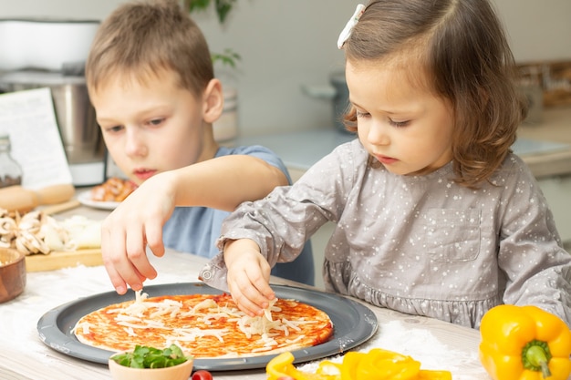 Bambina sveglia 2-4 in vestito grigio e ragazzo 7-10 in maglietta che cucina pizza insieme in cucina. Fratello e sorella che cucinano