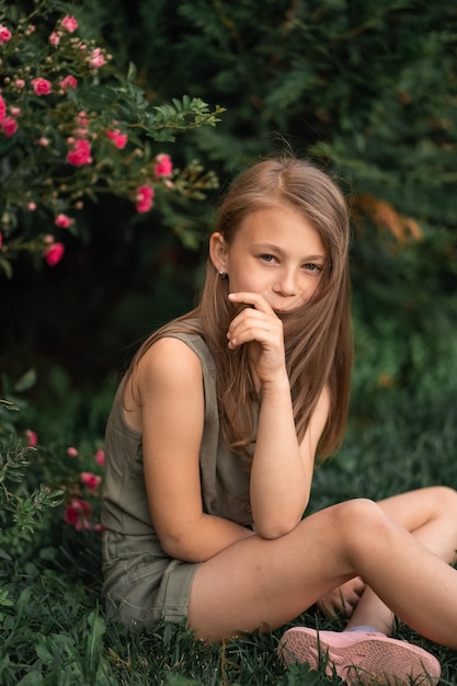 Bambina sveglia all'aperto che si siede vicino a un cespuglio di rose e si tocca i capelli