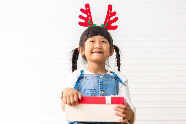 Bambina sveglia allegra di Natale e di feste felici che tengono il contenitore di regalo attuale al salone bianco.