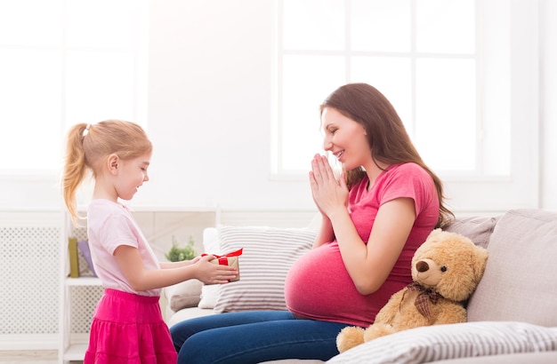 Bambina sveglia che dà il suo regalo di madre incinta a casa. Celebrazione della festa della mamma, figlia che si congratula con sua madre, copia dello spazio.