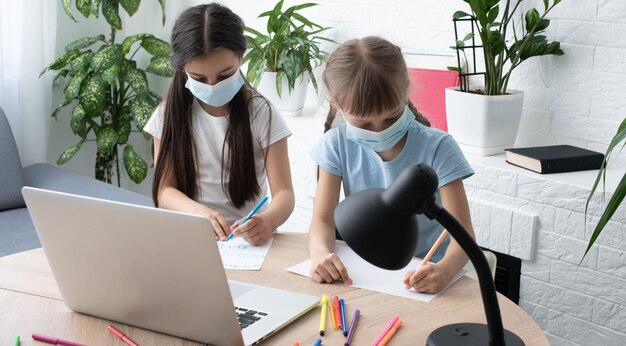 Bambina sveglia che indossa una maschera medica sul viso facendo apprendimento a distanza usando un laptop a casa in camera da letto. Una ragazza con una maschera protettiva sul viso tiene la testa in mano. Insegnamento a distanza