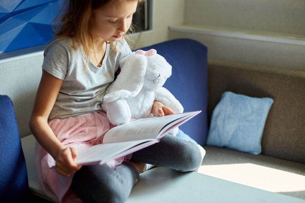 Bambina sveglia che mangia un libro con un coniglietto di peluche imbottito e sorride seduta sul divano in soggiorno. Resta a casa durante la pandemia di coronavirus covid-19