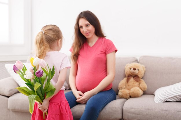 Bambina sveglia che nasconde i fiori da sua madre incinta dietro la schiena. Celebrazione della festa della mamma, figlia che si congratula con sua madre, copia dello spazio.