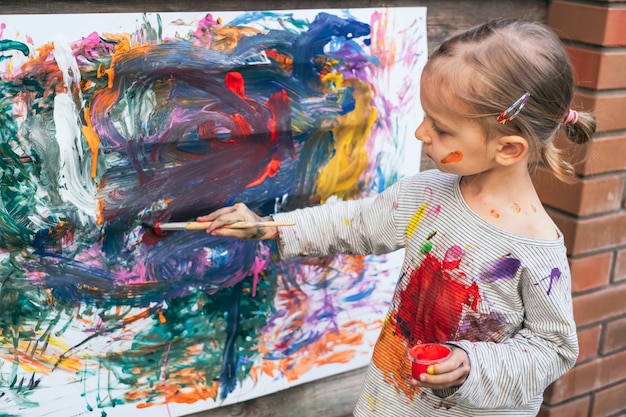 Bambina sveglia che si diverte, dipingendo con le dita con vari colori su carta sulla recinzione del cortile. Concetto di arte creativa per bambini.