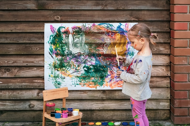 Bambina sveglia che si diverte, dipingendo con vari colori su carta sul recinto del cortile. Concetto di arte creativa per bambini.