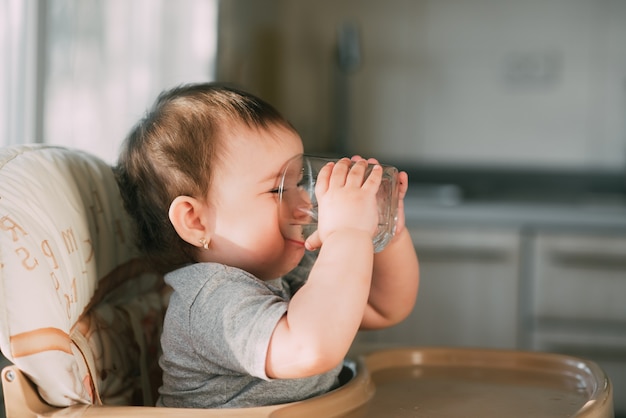 Bambina sveglia che si siede su un seggiolone in cucina e acqua potabile