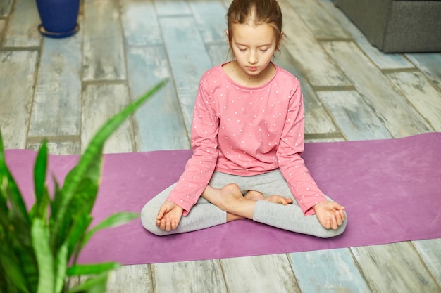 Bambina sveglia che si siede sul tappetino fitness in una posizione del loto mentre fa yoga a casa